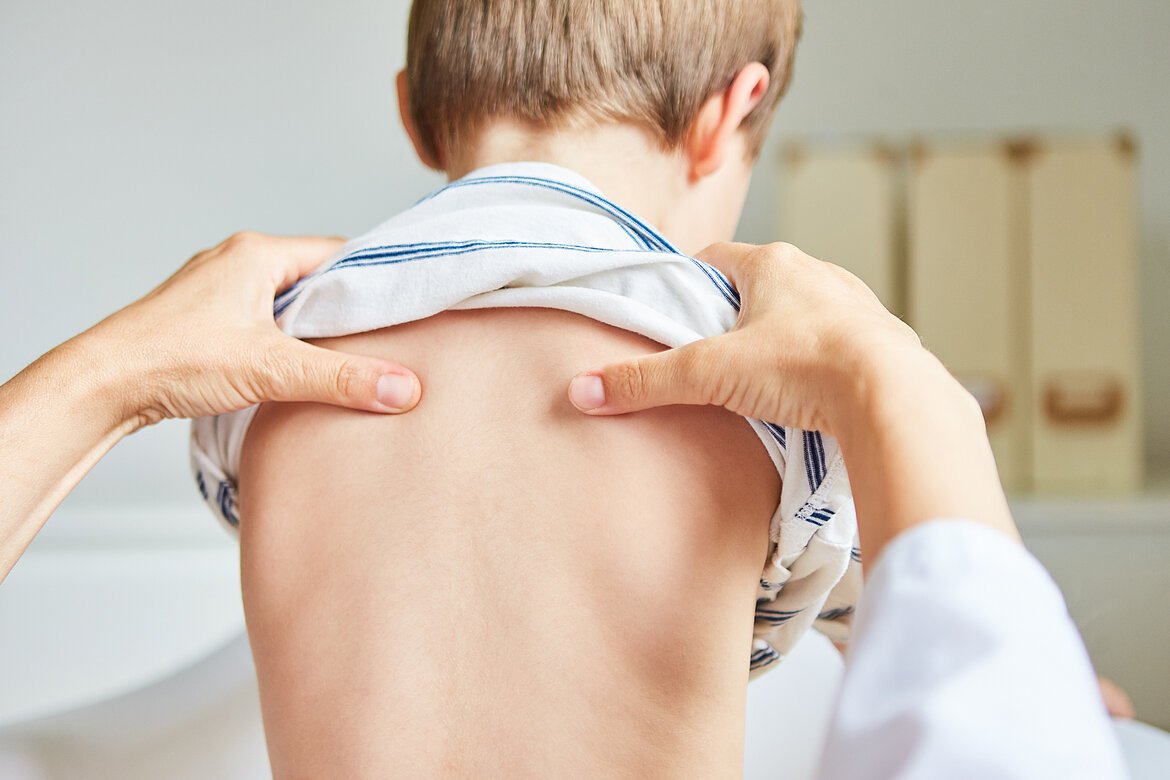 Männliches Kind mit hochgeschobenem T-Shirt in Rückenansicht. Osteopath druckt mit Daumen auf Schulterblätter.